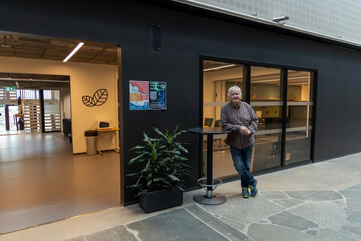 Hvithåret mann i rutete skjorte står foran et rom med vinduer inne på et universitetsbygg